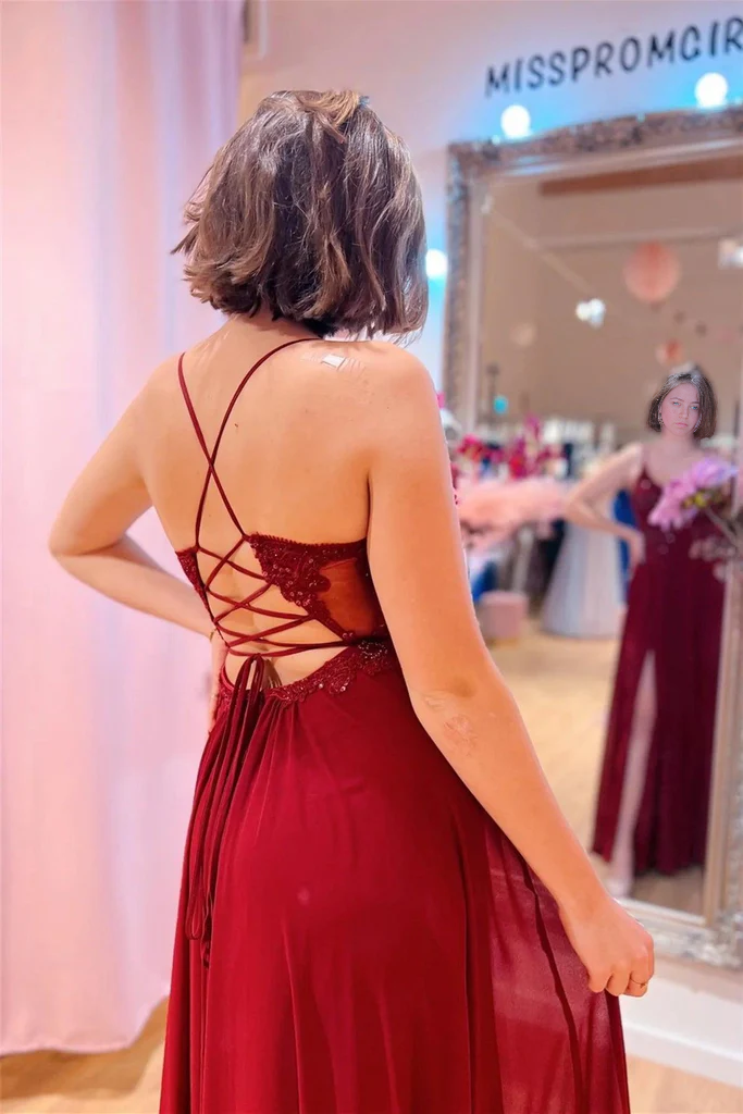 Full view of a woman wearing a long burgundy A-line prom dress with a V-neck and slit.