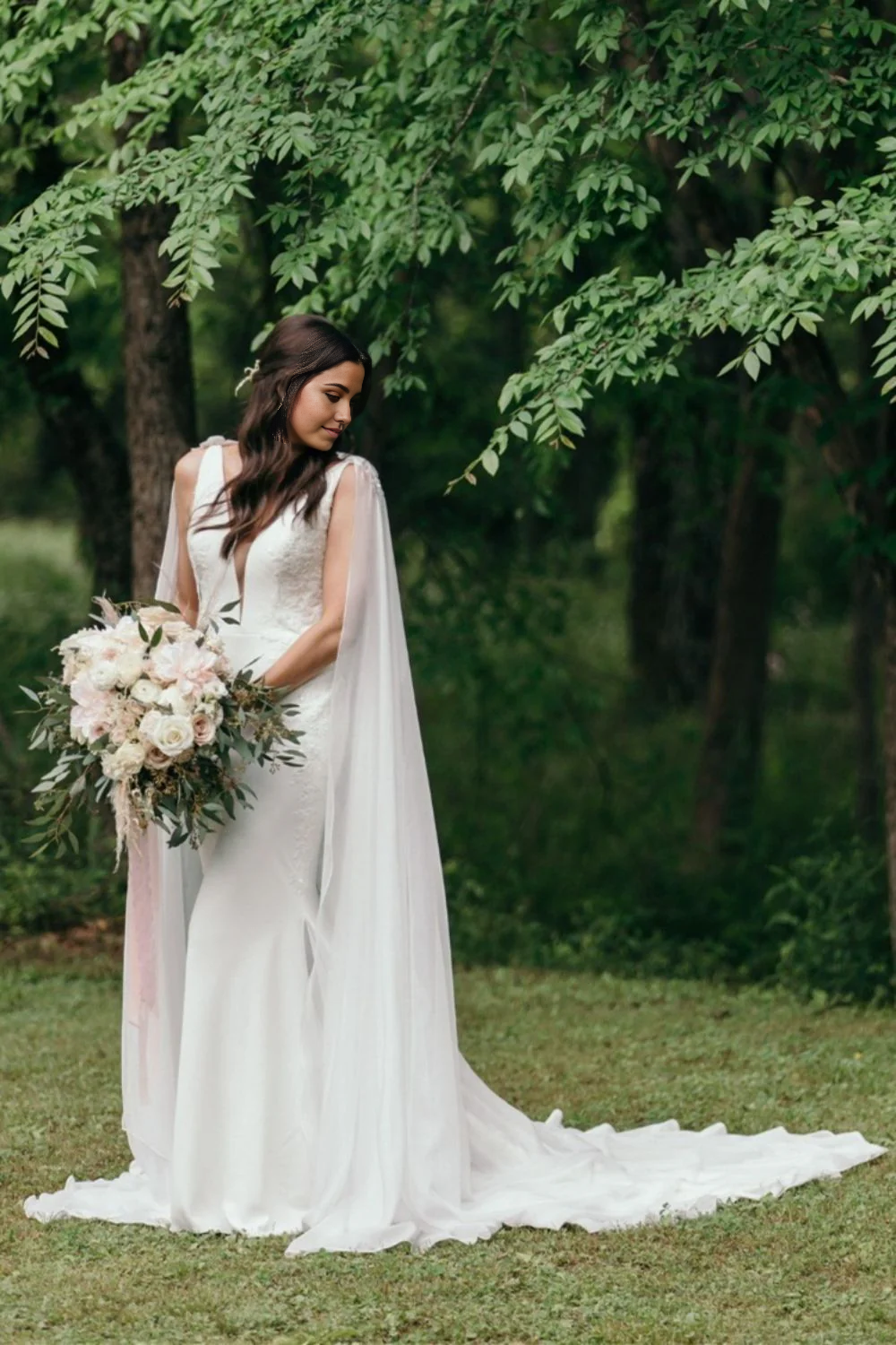 Model wearing an elegant white mermaid wedding dress with deep V-neck and flowing cape.