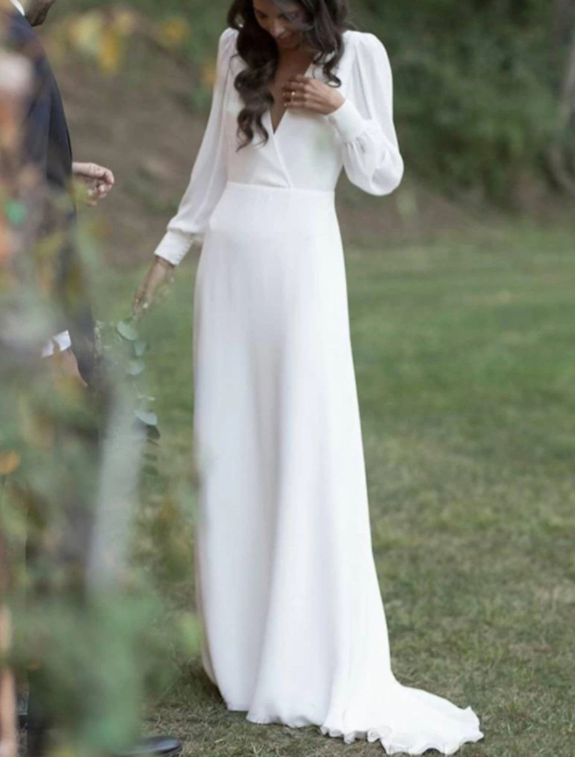 A bride wearing an elegant white long sleeve V-neck sheath wedding dress with an open back and sweep train.