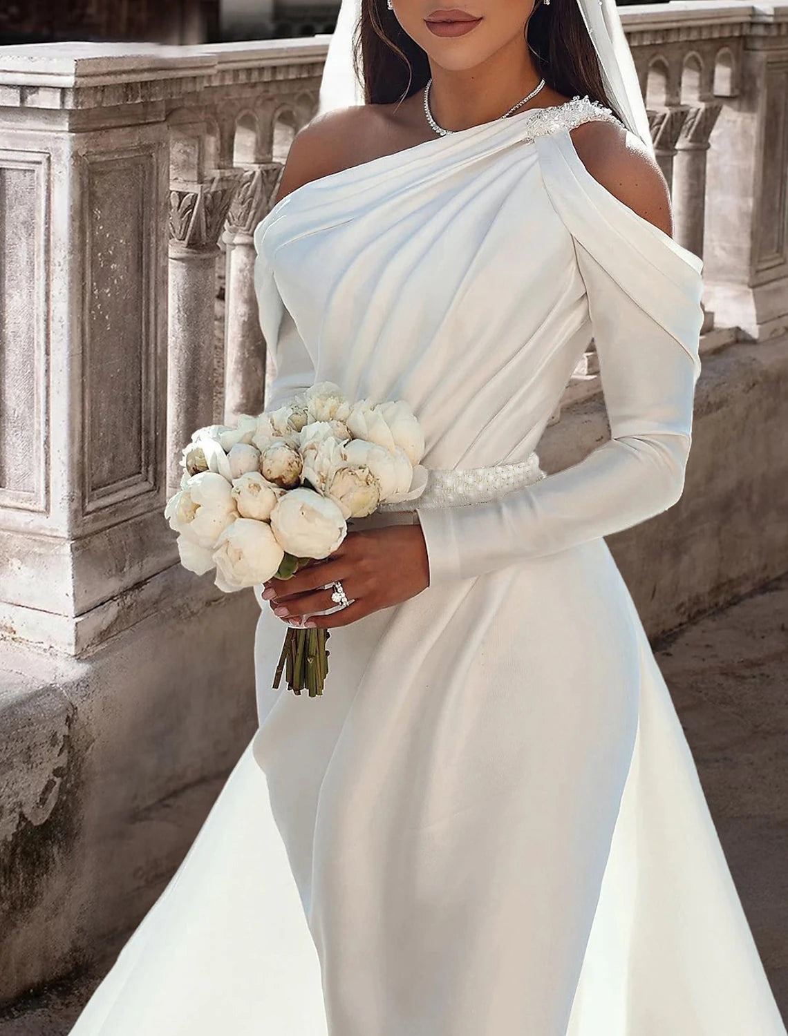Back view of a model in a two-piece one-shoulder long-sleeve satin bridal gown with a flowing train.