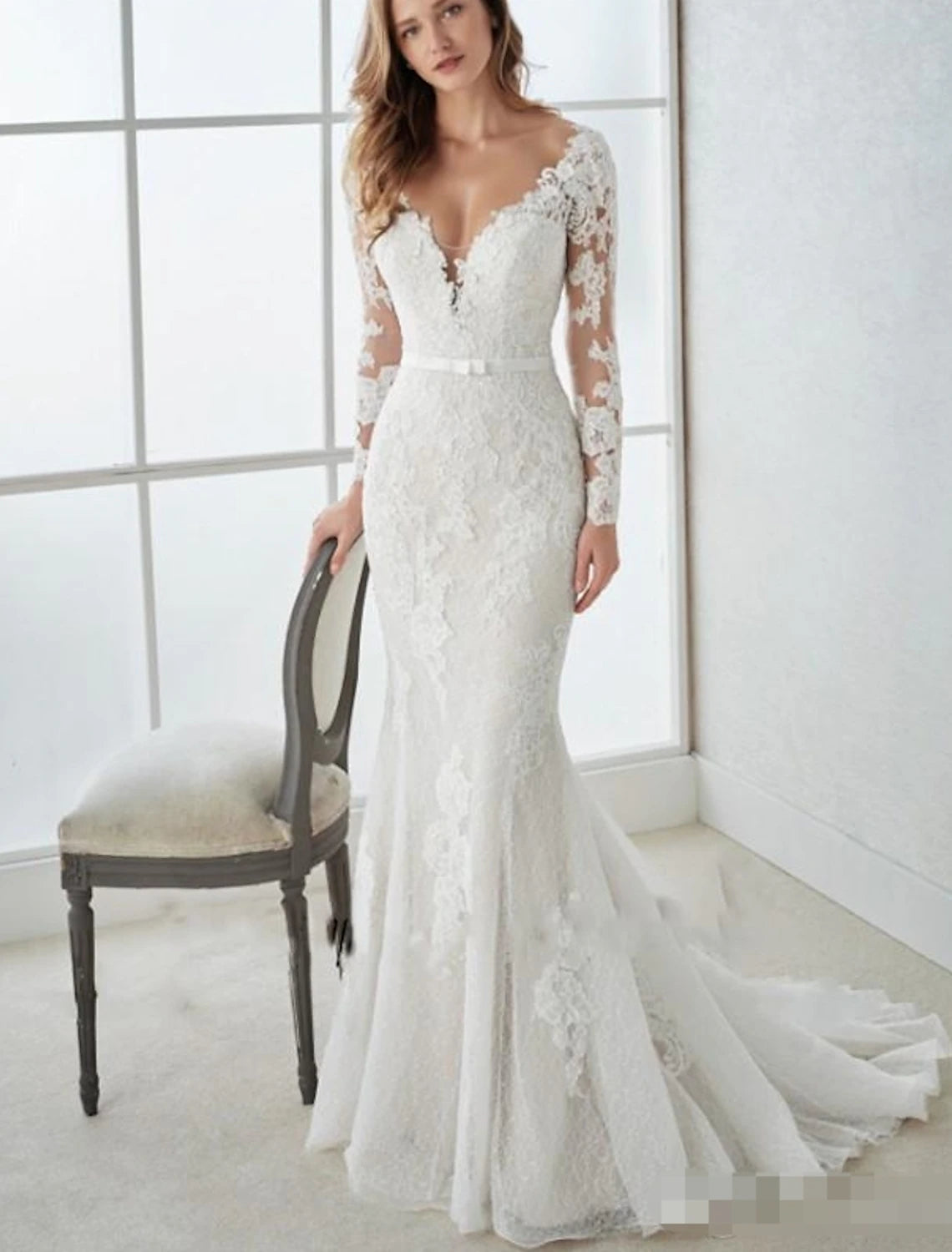 A woman wearing an elegant white mermaid V-neck long sleeve lace wedding dress with an open back and sweep train.