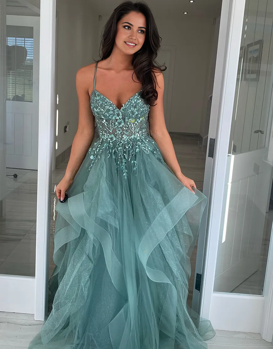 Model wearing a sparkly navy A-line prom dress with spaghetti straps, ruffled tulle, and floral details.